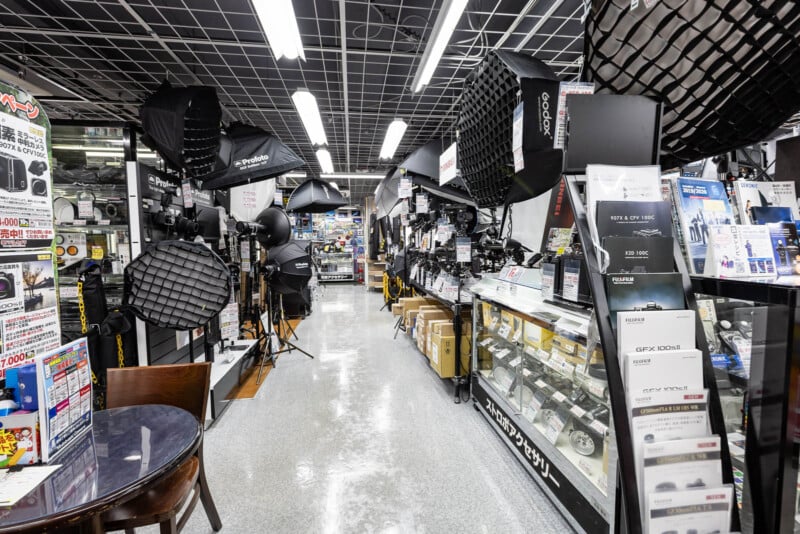 A well-lit camera store interior with photography equipment, studio lights, cameras, and accessories displayed on shelves and glass counters; posters and signs with Japanese text are visible.