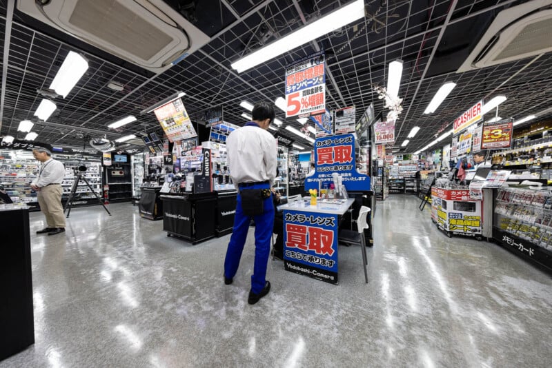 A wide-angle view of a brightly lit electronics store in Japan, with signs in Japanese, various gadgets on display, and a staff member in a white shirt and blue pants standing near a promotional sign.