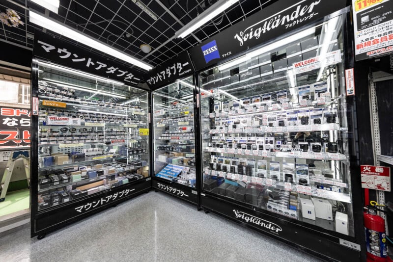 Glass display cases in a camera store showcase various camera lenses, adapters, and accessories. The shelves are filled with neatly arranged boxes and products, with Japanese and English signage above and around the displays.