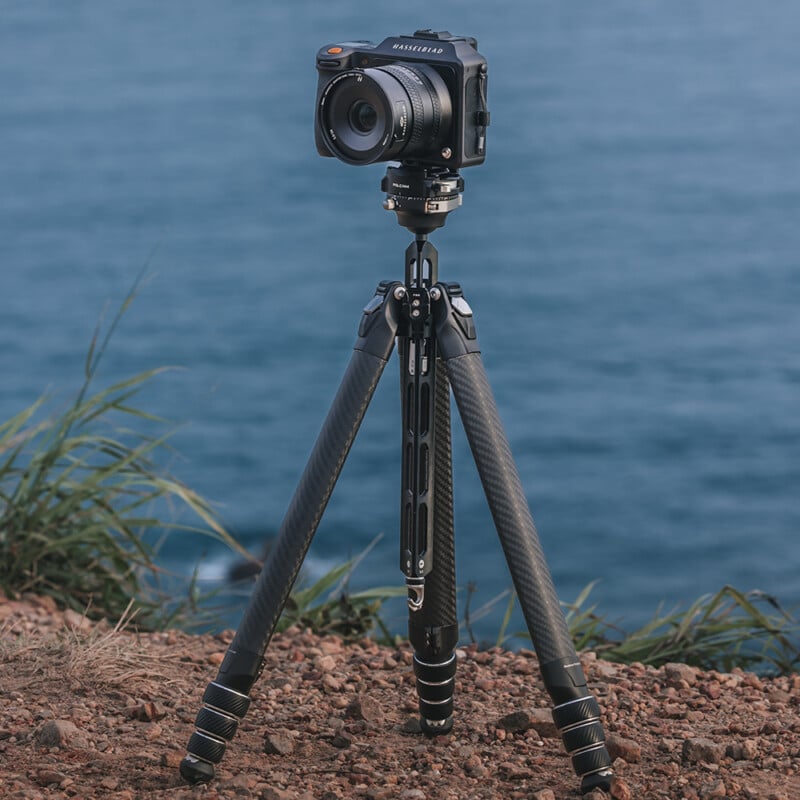 A professional camera is mounted on a sturdy tripod positioned on rocky terrain. The background showcases a serene body of water, suggesting the camera is set up for a scenic photography session. Green grass is visible on the edges of the rocky ground.