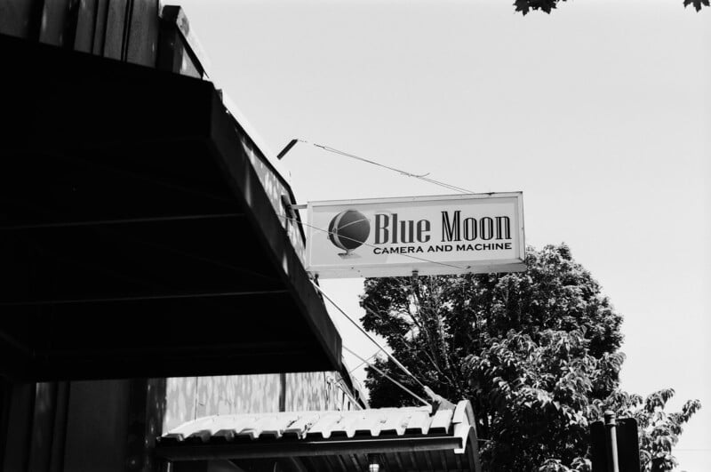 Black and white photo of a sign reading “Blue Moon Camera and Machine” with a globe logo, attached to the side of a building; tree branches and a clear sky are visible in the background.
