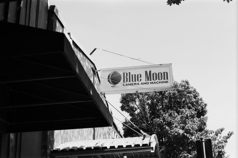 A black and white photo of a sign reading "Blue Moon Camera and Machine" attached to a building, with trees and part of the roof visible in the background.