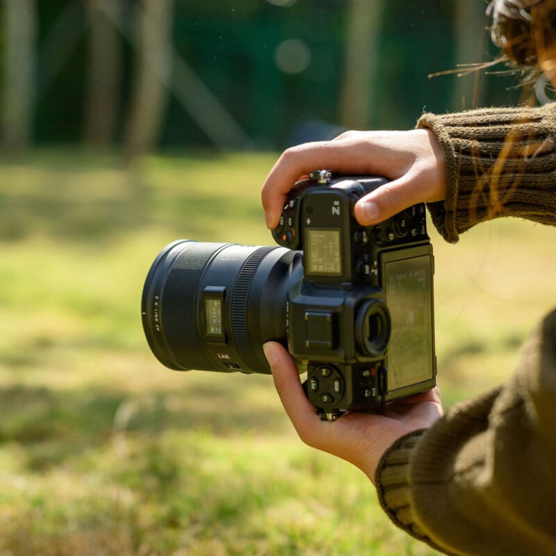 A person holding a digital camera with both hands, preparing to take a photo outdoors on a sunny day, with grass and trees blurred in the background.