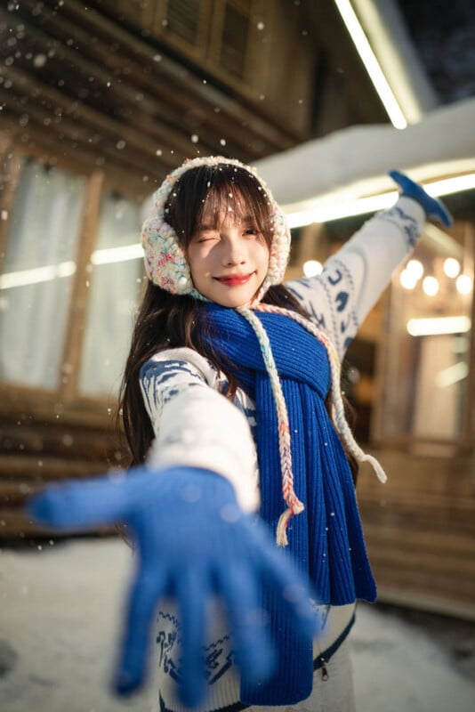 A smiling woman wearing earmuffs, a blue scarf, and blue gloves stands outside in falling snow with arms outstretched in front of a wooden cabin at night.
