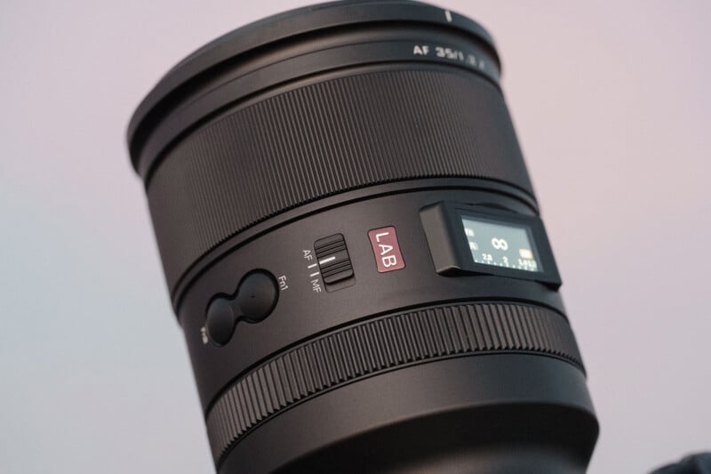 Close-up of a black camera lens with focus adjustment switches, a red "LAB" label, and a digital display showing infinity focus and aperture settings. The background is softly blurred in light pastel tones.