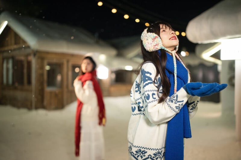 Two women stand outside in a snowy village at night. The woman in front wears earmuffs, blue scarf, blue gloves, and a patterned sweater, looking up and smiling with hands outstretched. Another woman stands in the background.