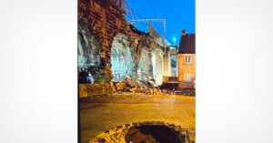 A damaged brick railway viaduct with a collapsed section, scattered debris, and a large hole in the cobblestone street below, illuminated by streetlights at dusk.