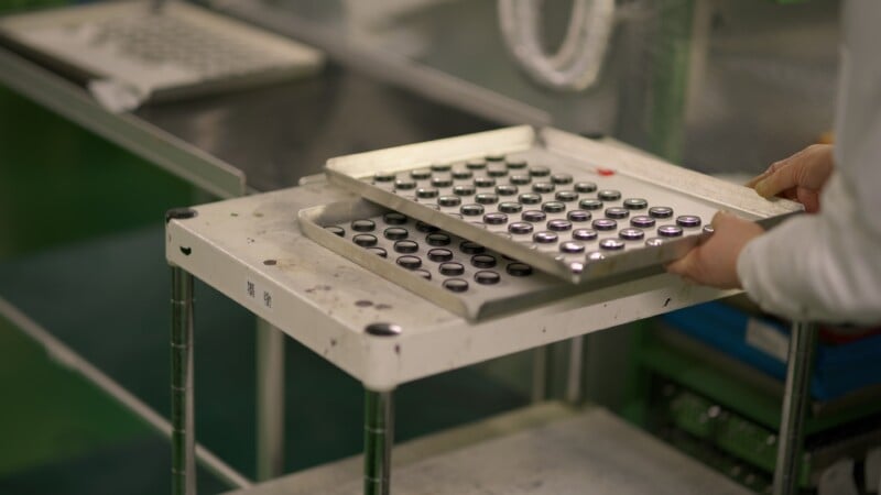 A person in a lab coat handles a metal tray filled with small cylindrical objects, possibly batteries, on a cart in an industrial or laboratory setting.
