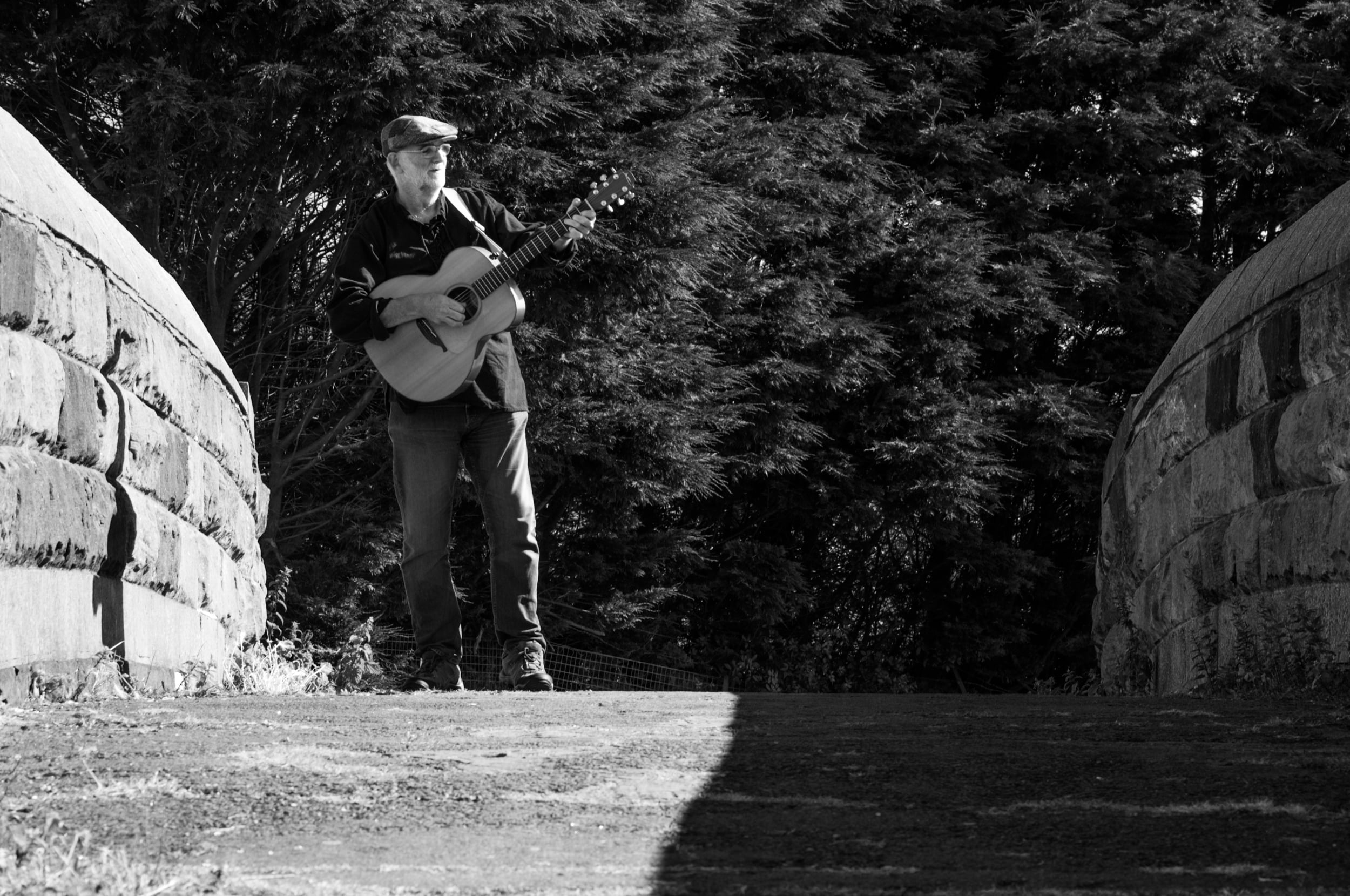 A man wearing a hat plays an acoustic guitar while standing on a stone bridge. Tall trees fill the background, and the scene is in black and white with sunlight casting shadows on the ground.