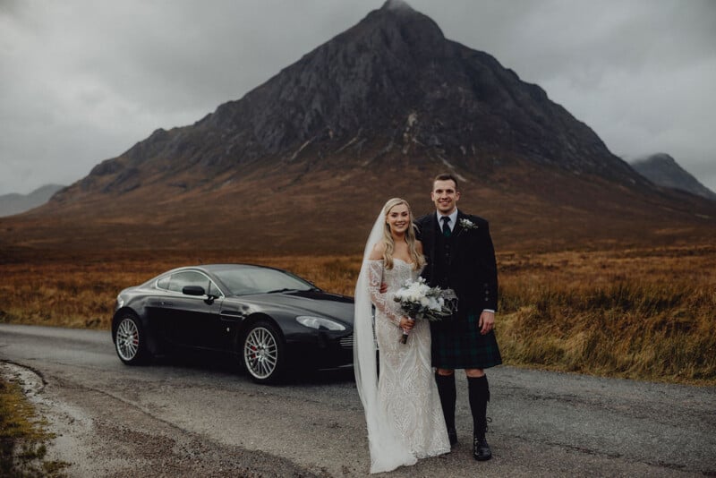A bride and groom stand smiling on a rural road in front of a black sports car, with a dramatic mountain and cloudy sky in the background. The bride holds a bouquet and the groom wears a traditional kilt.