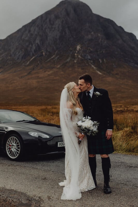 A bride and groom kiss in front of a black car on a rural road, with a mountain in the background. The bride wears a white gown and holds a bouquet; the groom wears a kilt and jacket. The scene appears overcast and romantic.