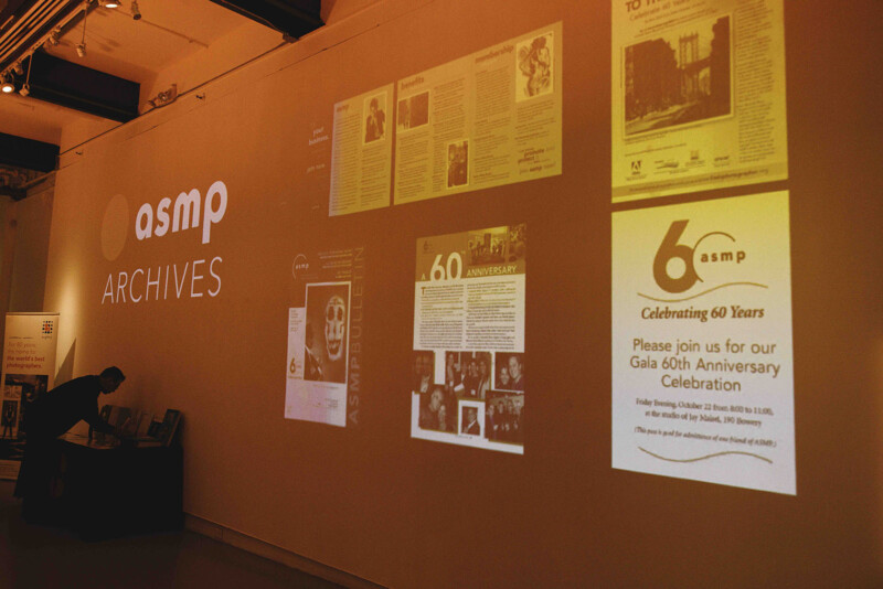 A wall displays "asmp ARCHIVES" alongside projected images of documents and flyers, including one inviting guests to a 60th anniversary celebration. A person stands at a table in the dimly lit room.