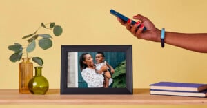 A person takes a photo of a framed picture showing a smiling woman holding a baby. The frame sits on a wooden shelf with books, green glass vases, and leafy branches, against a yellow background.
