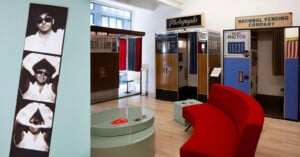 A photo strip with three black-and-white images of a man wearing sunglasses is displayed on a wall next to a room with vintage photo booths, a red curved couch, and a round table.
