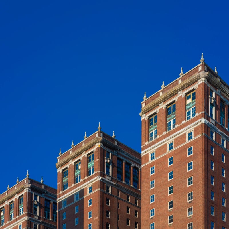 Three tall, red-brick buildings with decorative white trim stand against a clear, vibrant blue sky. The buildings have multiple windows and ornate architectural details along the rooflines.