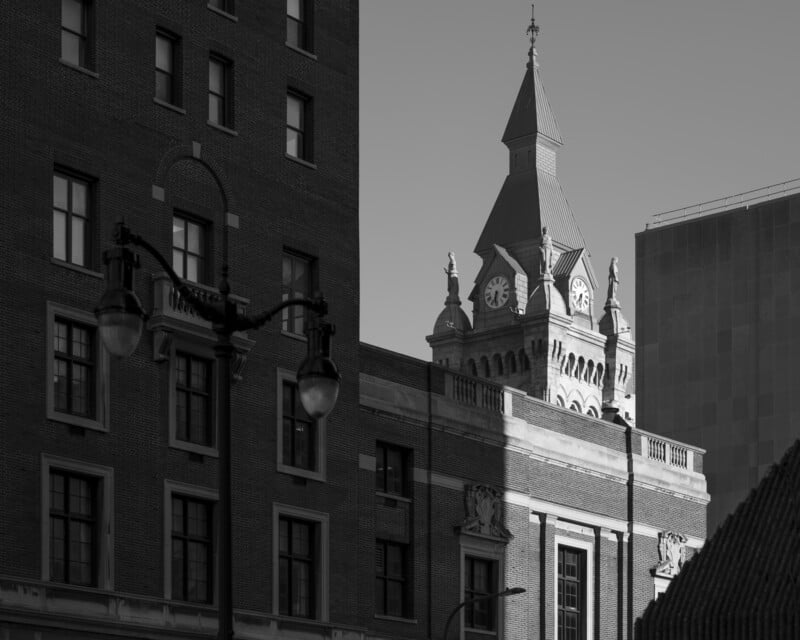 Black and white photo of a historic building with a clock tower, framed by modern buildings and a streetlamp in the foreground under clear skies.