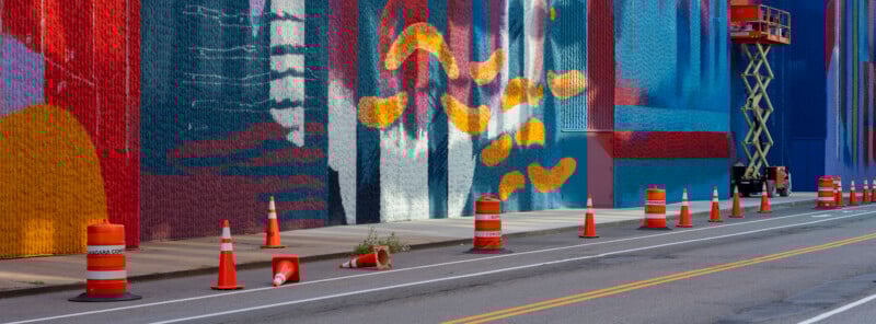 A colorful mural covers a tall wall along a street lined with orange traffic cones. An orange lift is raised near the wall, and one cone is knocked over with a small plant growing next to it.