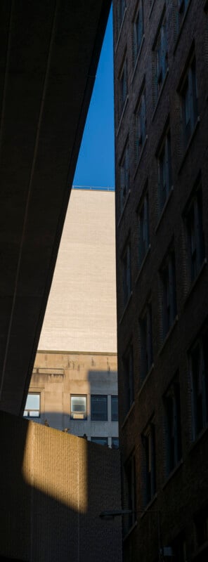 View looking up between two dark, tall buildings, with a patch of bright sunlight illuminating parts of a wall and a clear blue sky visible at the top.