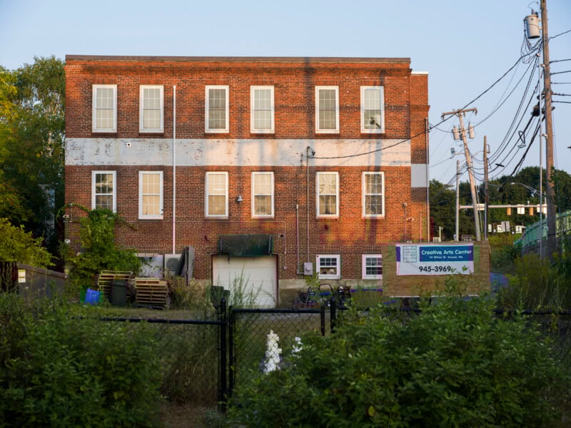 A three-story brick building with white trim, surrounded by greenery and utility poles. A sign in front reads "Creative Arts Center" with a phone number. Various items and pallets are stacked by the building.