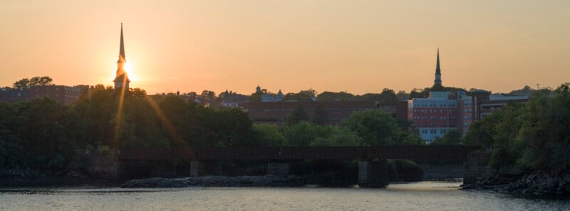Sun sets behind a cityscape with church steeples, a mix of trees, brick buildings, and a railroad bridge crossing a calm river in the foreground.