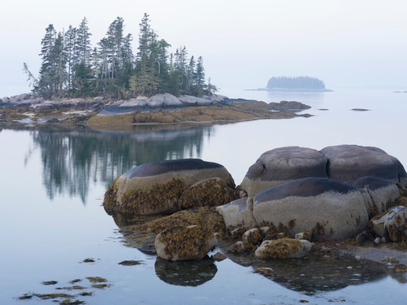 A calm coastal scene features large, rounded rocks in the foreground and a small wooded island reflected in the still water, with another distant island visible through light mist.