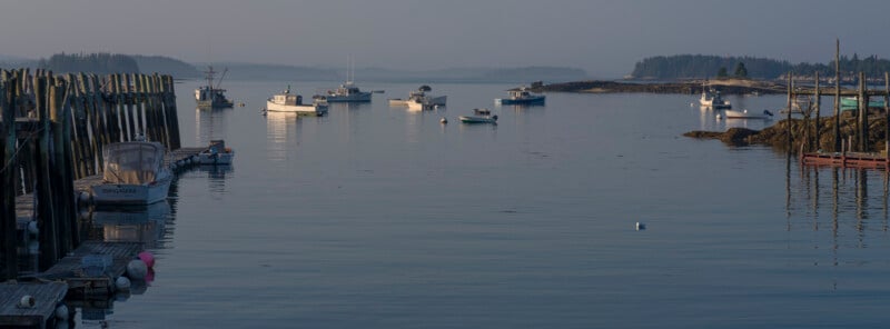 Boats float on calm water near a wooden dock and rocky shore, with soft light and a hazy sky. Small islands and forested land are visible in the distance.