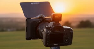A Canon DSLR camera mounted on a tripod faces a sunset, equipped with a large ND filter and hood. The background is a blurred landscape of green fields and a glowing orange sky.