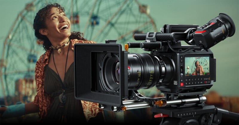 A woman with curly hair looks upward and smiles joyfully. In the foreground, there is a large professional camera with a monitor displaying her image. A Ferris wheel is blurred in the background.