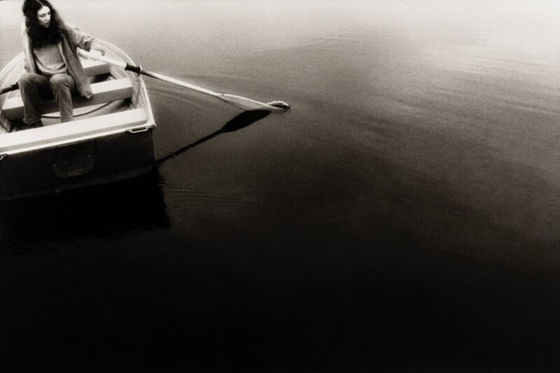 A person sits alone in a small rowboat on calm, dark water, holding one oar. The boat is positioned in the lower left corner, with most of the image showing the still, reflective surface of the water.