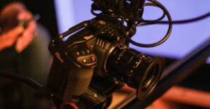 A close-up of a professional video camera with cables attached, set up on a tripod against a blurred background.