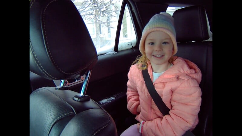 A young child wearing a pink jacket and a light-colored knit hat sits in the back seat of a car, smiling at the camera with a seatbelt fastened. Snowy scenery is visible through the window.