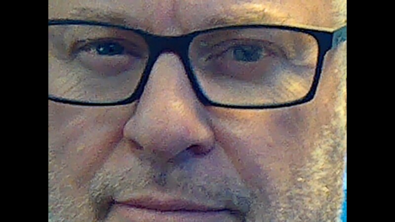 A close-up image of a middle-aged person’s face wearing black-framed glasses, with stubble, light skin, and blue eyes looking slightly to the left. The background is out of focus.