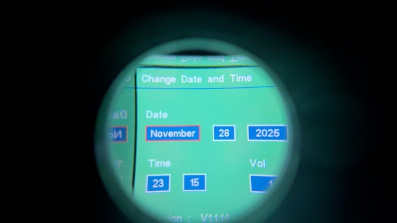 A digital screen viewed through a circular lens displays a date and time setting: November 28, 2025, with time options shown as 23 and 15. The interface has a green background and blue buttons.