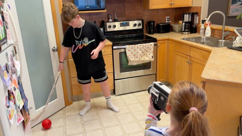 A boy in a black shirt and shorts opens a door with a stick in a kitchen, while another child with light brown hair films him using a vintage video camera. The kitchen has wooden cabinets and appliances.