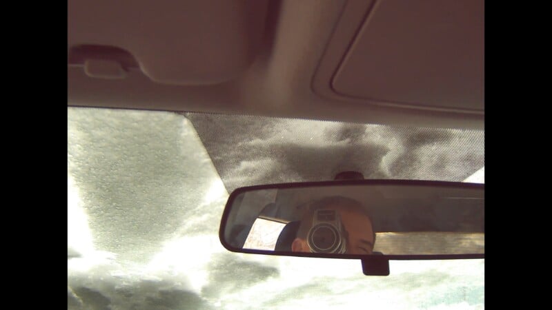 View from inside a car looking up through the windshield at cloudy sky, with a rearview mirror reflecting a person holding a camera taking a picture.