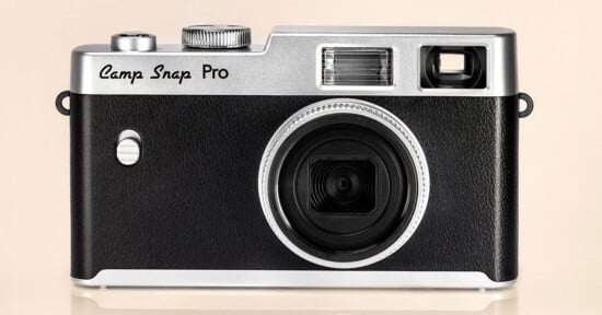 A black and silver retro-style digital camera labeled "Camp Snap Pro," shown from the front against a beige background. The camera has a large lens, flash, and viewfinder.