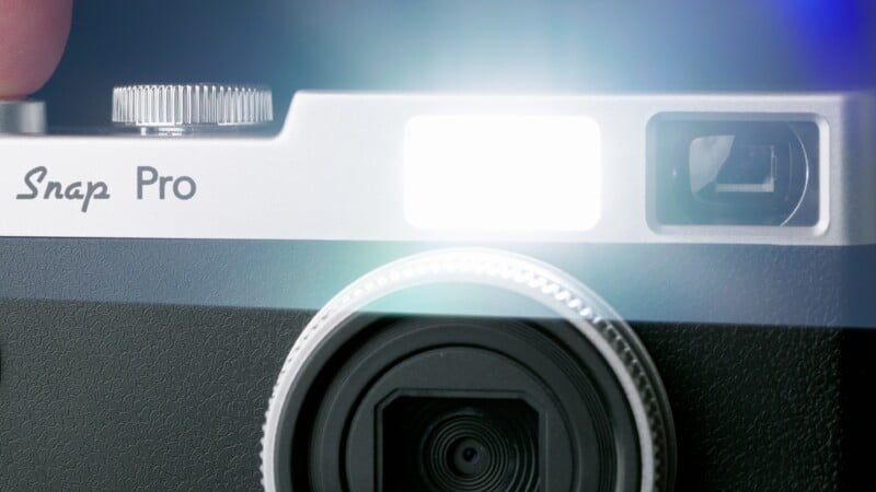 Close-up of a camera’s top front, showing the lens, viewfinder, and a bright flash firing. The camera has a silver and black body, with the text “Snap Pro” visible on the metallic part.