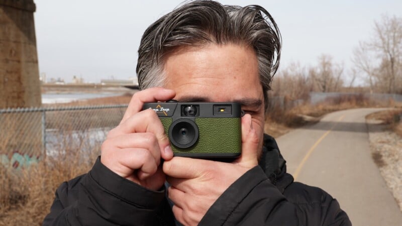 A person with dark hair is outdoors, holding a green film camera up to their face, ready to take a picture. They are wearing a black jacket, and a pathway, fence, and leafless trees are visible in the background.
