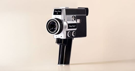 A vintage black and silver Super 8 film camera with a large lens and a pistol grip handle, set against a plain, light beige background.