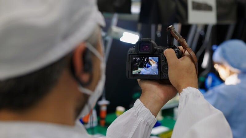 A person in protective clothing and a hairnet is holding a camera, photographing or filming another person in similar attire who appears on the camera’s screen, likely in a laboratory or medical setting.