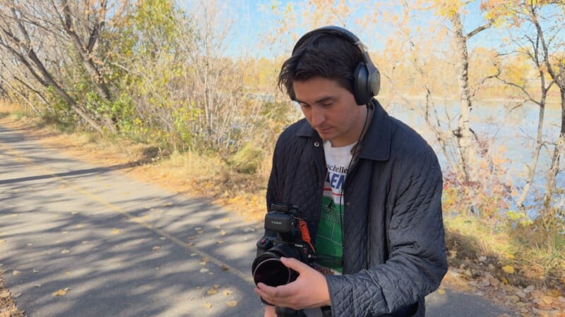 A person wearing headphones and a dark jacket stands on a sunlit path lined with autumn trees, holding and looking at a camera and microphone setup.