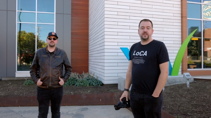 Two men stand outside a modern building. One wears a black leather jacket, sunglasses, and cap. The other wears a black "LoCA" t-shirt and holds a camera. The background features white and wood paneling with green and blue accents.