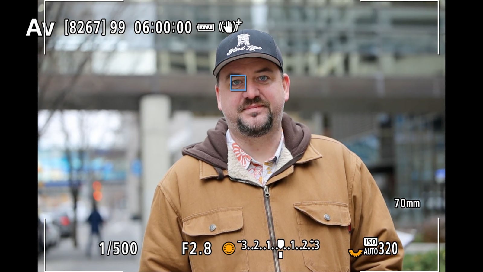 A man in a brown jacket and black cap stands outdoors, facing the camera. The image appears through a camera viewfinder, displaying settings like ISO 320, F2.8, 1/500 shutter speed, and focus on his face.
