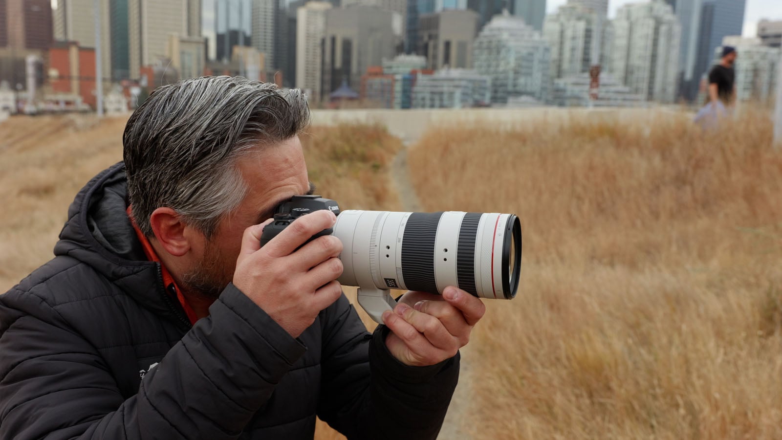 A man with gray hair and a beard, wearing a black jacket, is holding a camera with a large telephoto lens, taking a photo in a grassy field with a city skyline in the background.