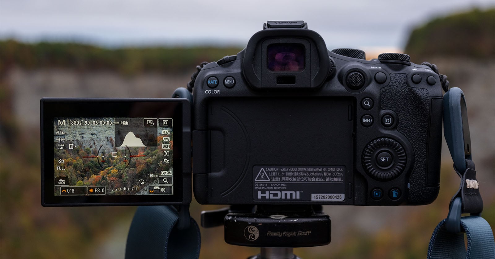 A digital camera on a tripod shows a rocky landscape with autumn trees on its LCD screen. The camera's buttons, dials, and HDMI port are visible, with the live view screen tilted to the side.