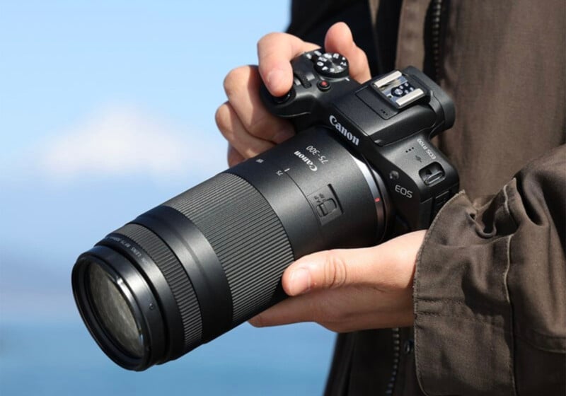 A person holding a Canon EOS camera with a large telephoto lens, wearing a brown jacket, with a blurred outdoor background.