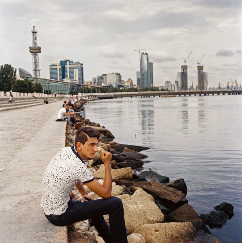 A young man in a white shirt sits on rocks by the water, resting his chin on his hand. Other people sit along the waterfront promenade. City buildings and cranes are visible in the background under a cloudy sky.