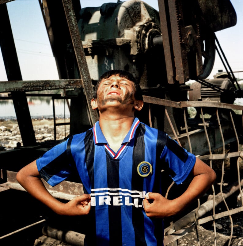 A boy wearing a blue and black Inter Milan soccer jersey, his face and arms dirty, stands outdoors in front of industrial machinery, squinting upwards with a determined expression and hands gripping his shirt.