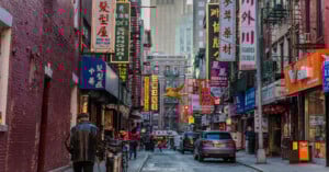 A busy street in Chinatown with colorful shop signs in Chinese, people walking, parked bikes, and cars. Tall buildings rise in the background, and a golden dragon decoration hangs above the street.