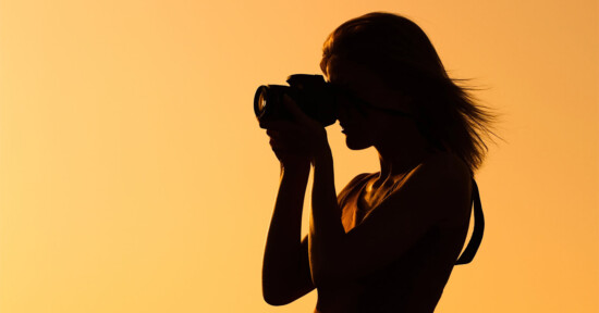 Silhouette of a person holding a camera and taking a photo against a bright orange-yellow background, likely at sunset or sunrise.